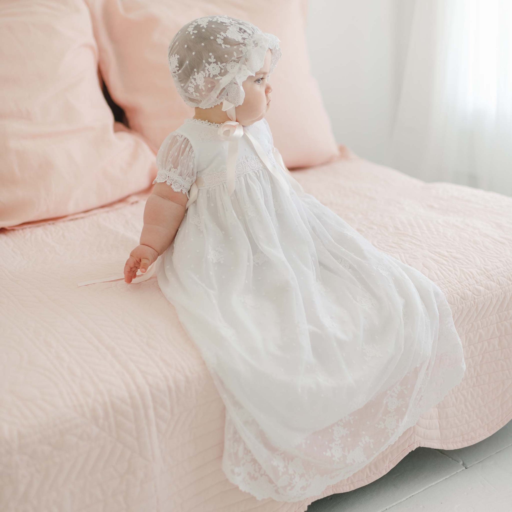 A baby dressed in a long white Melissa Christening Gown & Bonnet adorned with Venice lace trim is sitting on a pink bed. The baby is facing to the side, and the gown drapes elegantly over the bed's edge. The bed, with its pink pillows and quilted bedspread, appears softly lit in the cozy room.
