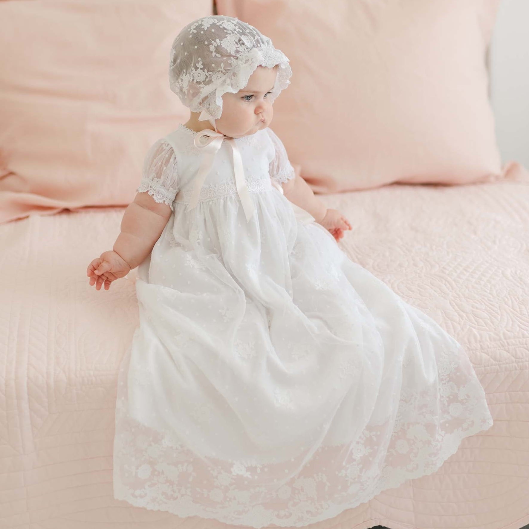 A baby wearing a Melissa Christening Gown & Bonnet is sitting on a bed with a light pink quilt and pillows. The baby is looking toward the side, and the room has a soft, pastel color scheme.