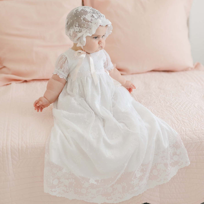 A baby wearing a Melissa Christening Gown & Bonnet is sitting on a bed with a light pink quilt and pillows. The baby is looking toward the side, and the room has a soft, pastel color scheme.