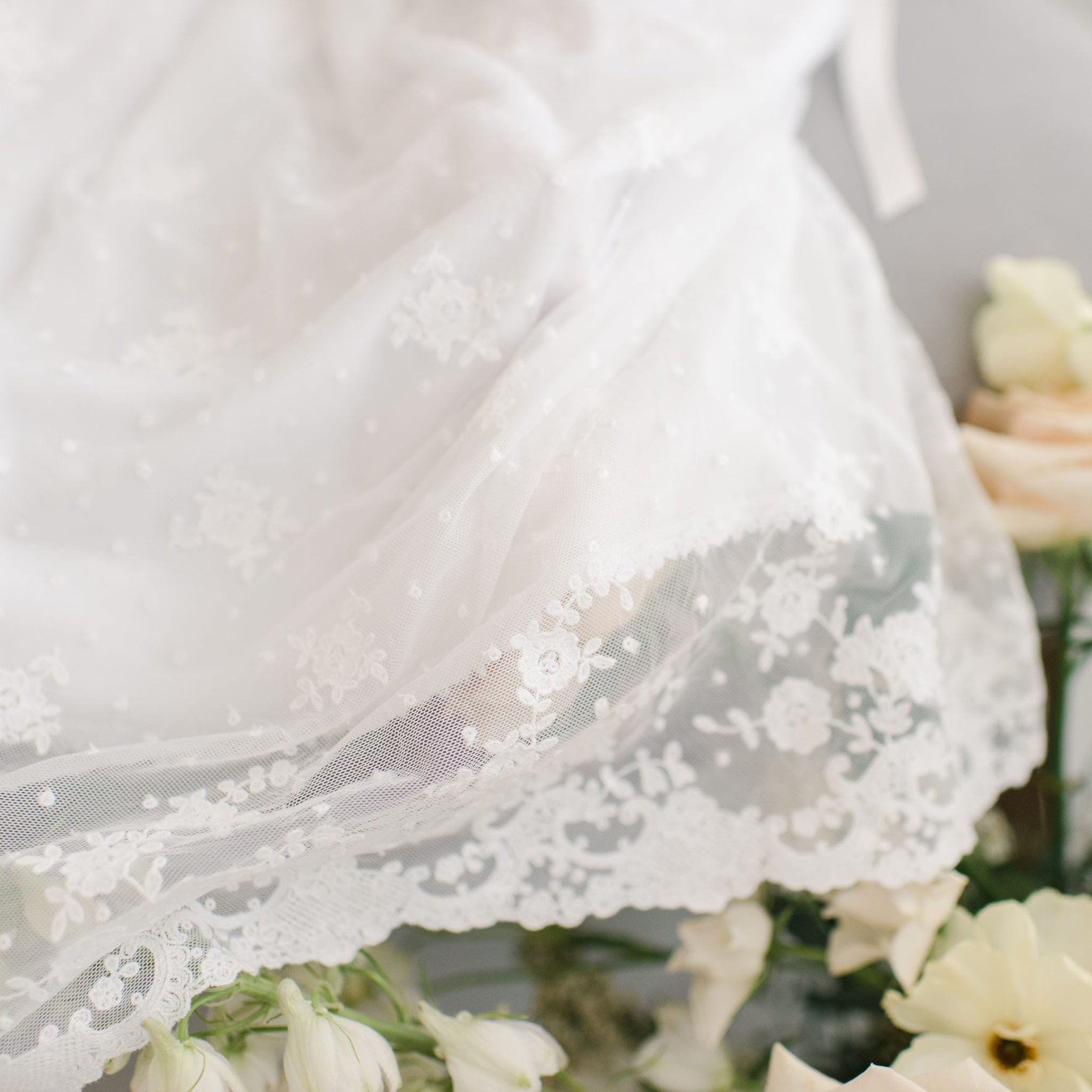 A close-up of delicate white lace fabric with floral and dotted patterns, possibly from a Melissa Christening Gown & Bonnet, draped over light-colored flowers. The intricate lace detail, featuring Venice lace trim, prominently showcases the craftsmanship.
