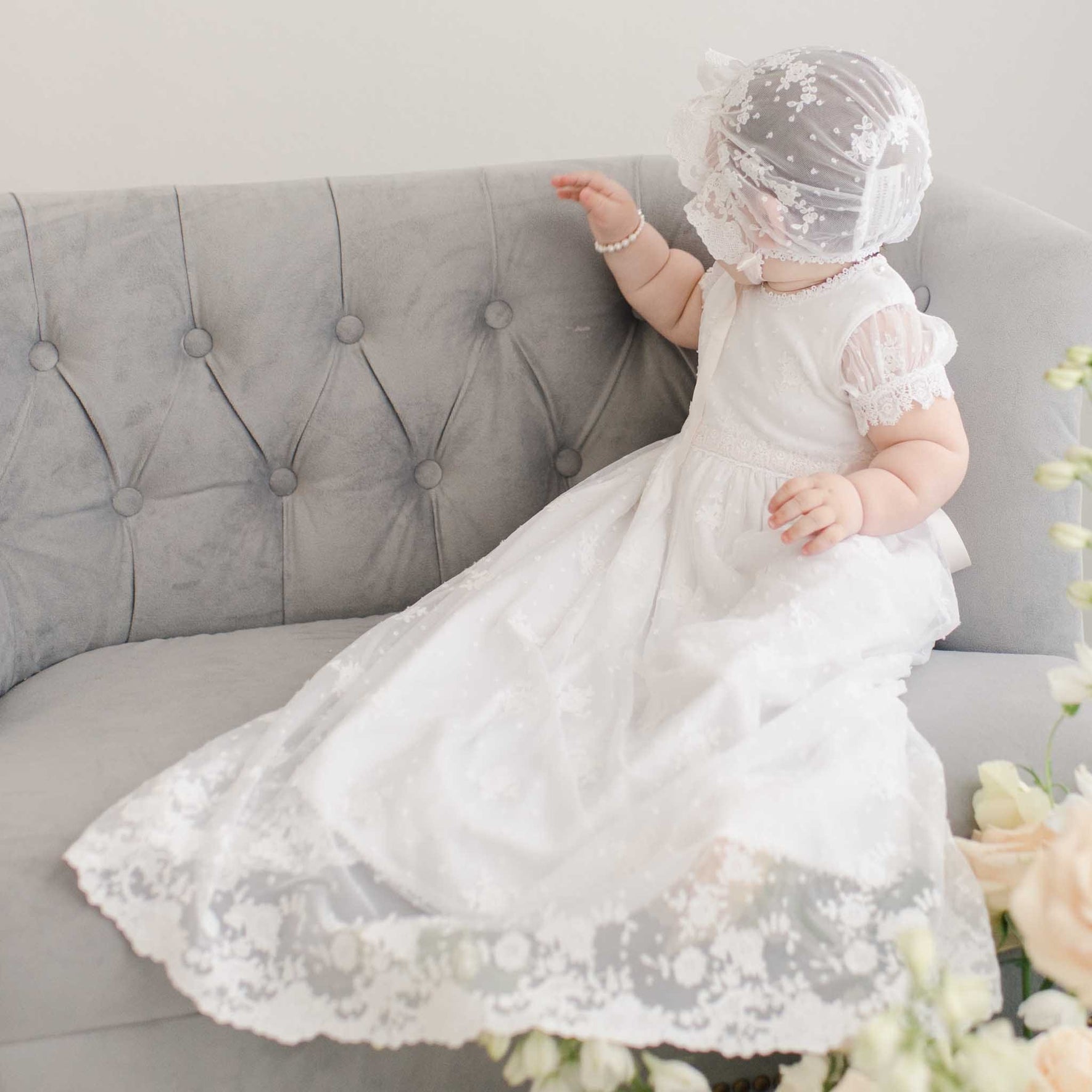 A baby wearing the Melissa Christening Gown & Bonnet adorned with Venice lace trim sits on a gray tufted sofa, facing away from the camera. Handmade in the USA, the outfit is completed with a pearl bracelet and surrounded by a soft floral arrangement.