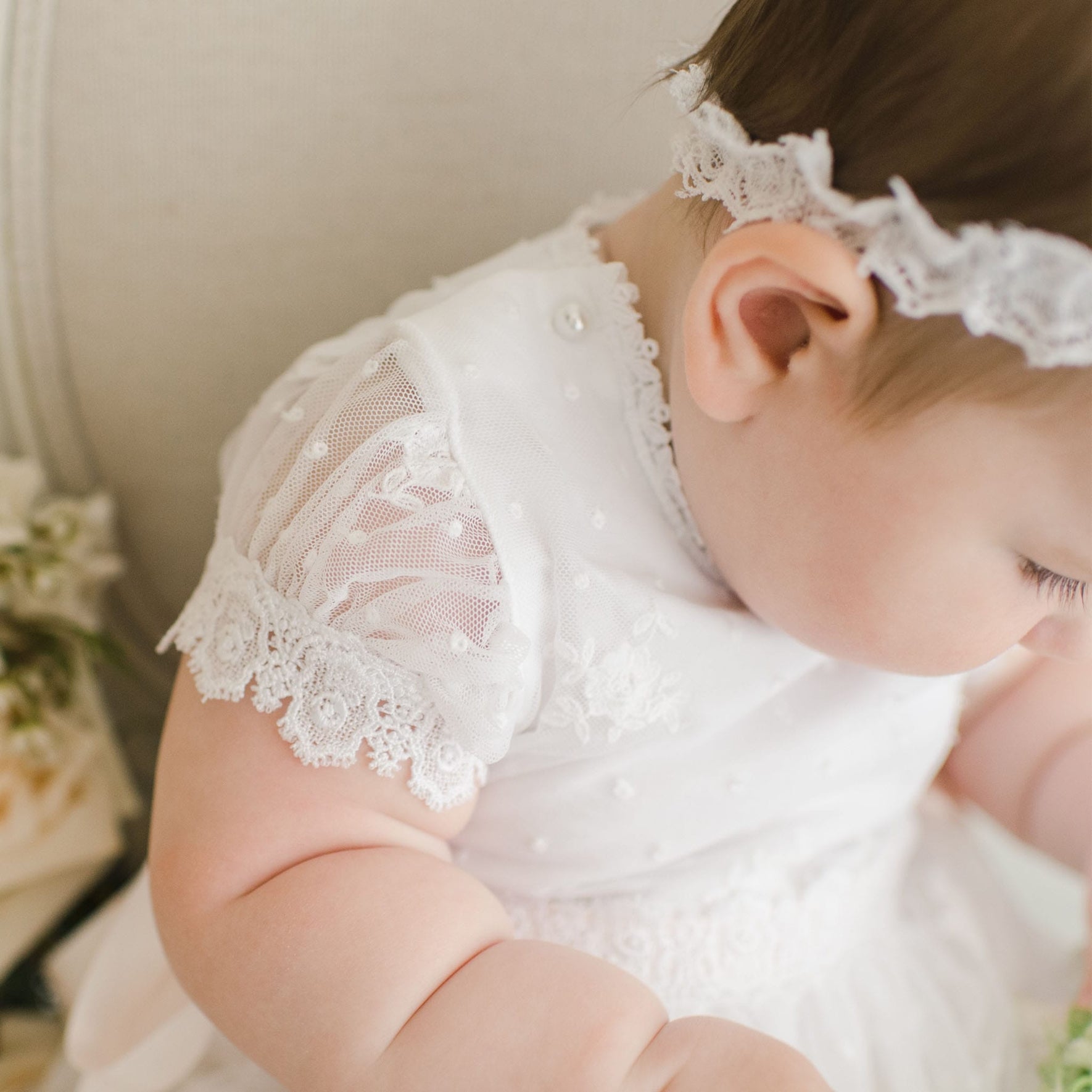 A baby wearing a Melissa Christening Gown & Bonnet sits on a light-colored chair. The gown features intricate lace detailing on the sleeves and pearls. The baby looks downward and is positioned slightly to the side. Flowers are in the background, highlighting its handmade quality from the USA.