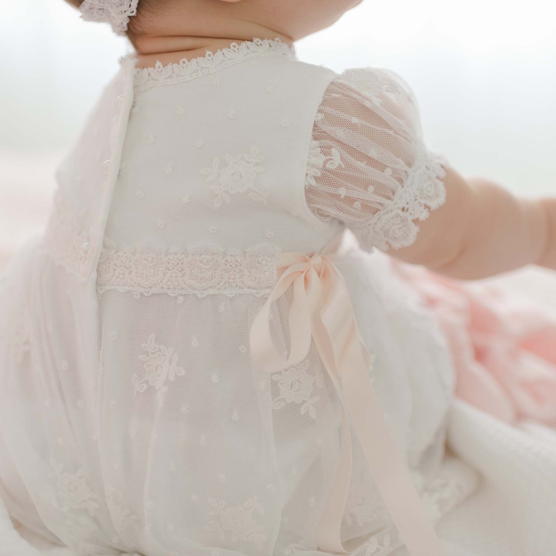 A baby in a delicate white Melissa Christening Gown & Bonnet with lace details and a pink ribbon sits with its back to the camera. Handmade in the USA, the gown features sheer short sleeves, Venice lace trim, and embroidered patterns throughout. The baby is on a soft surface with a light-colored background.