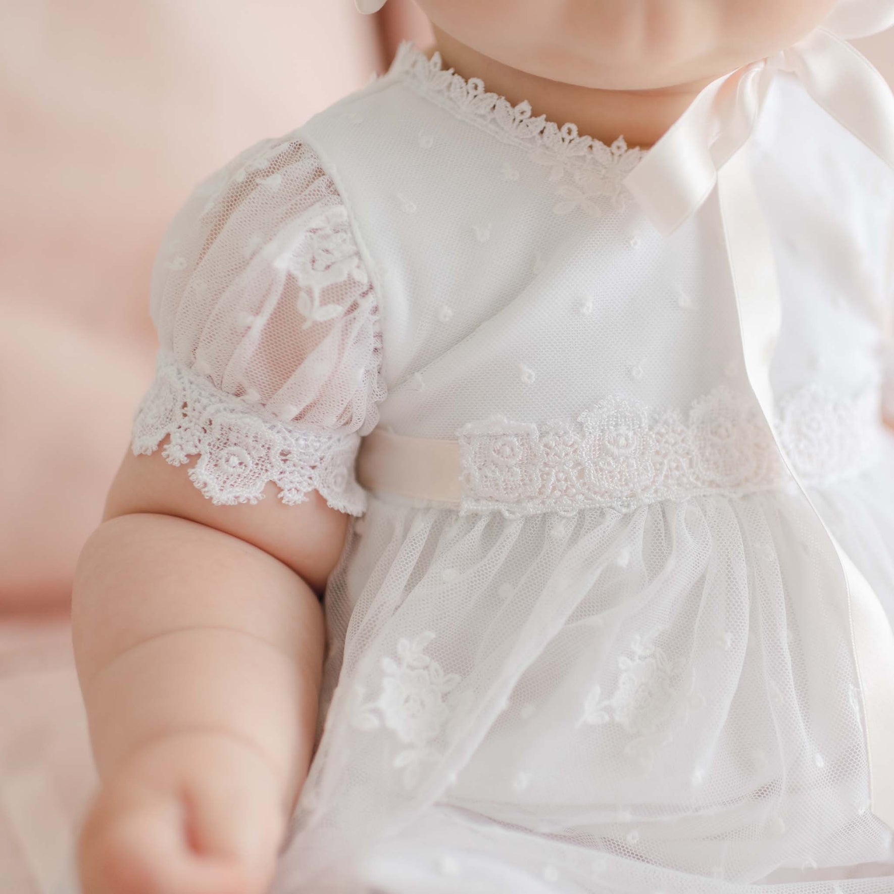 Close-up of a baby wearing a delicate white lace dress. The Melissa Christening Gown & Bonnet has short sleeves with intricate Venice lace trim, a satin ribbon bow at the neck, and a floral lace design on the bodice and skirt. Handmade in the USA, the baby's arm is slightly extended in the foreground.