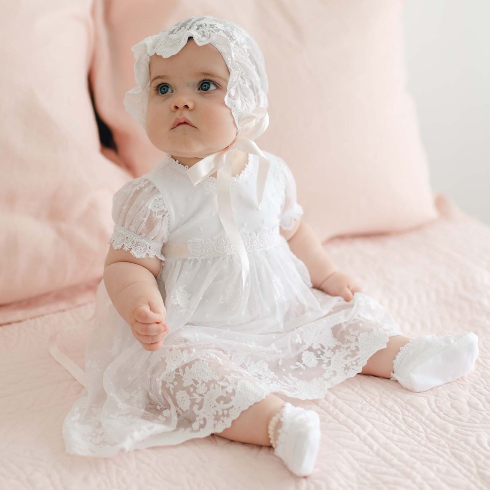 A baby dressed in a handmade Melissa Christening Romper Dress with matching bonnet sits on a bed with light pink bedding and pillows. The baby is looking slightly upward with a calm expression.
