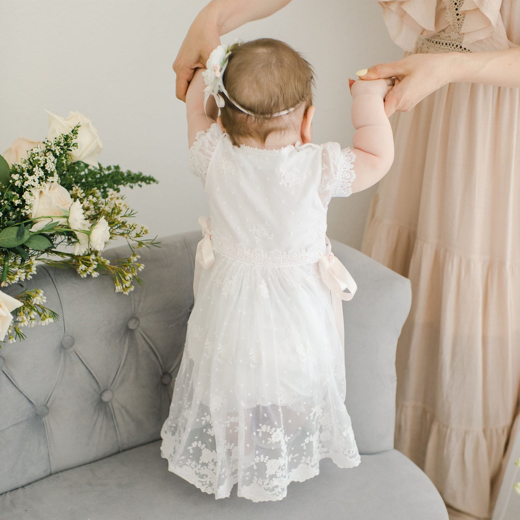 A baby wearing a Melissa Christening Romper Dress stands on a gray upholstered chair, holding onto the hands of an adult. White roses with greenery are arranged next to her.