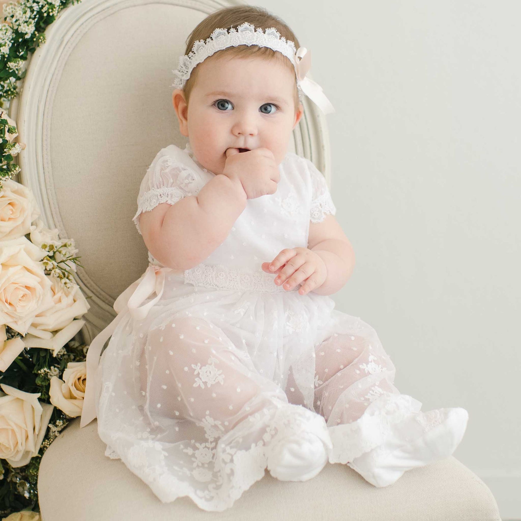 A baby in a Melissa Christening Romper Dress and headband sits on a cream-colored chair with a bouquet of roses next to her. The baby has one hand in her mouth and is looking at the camera.