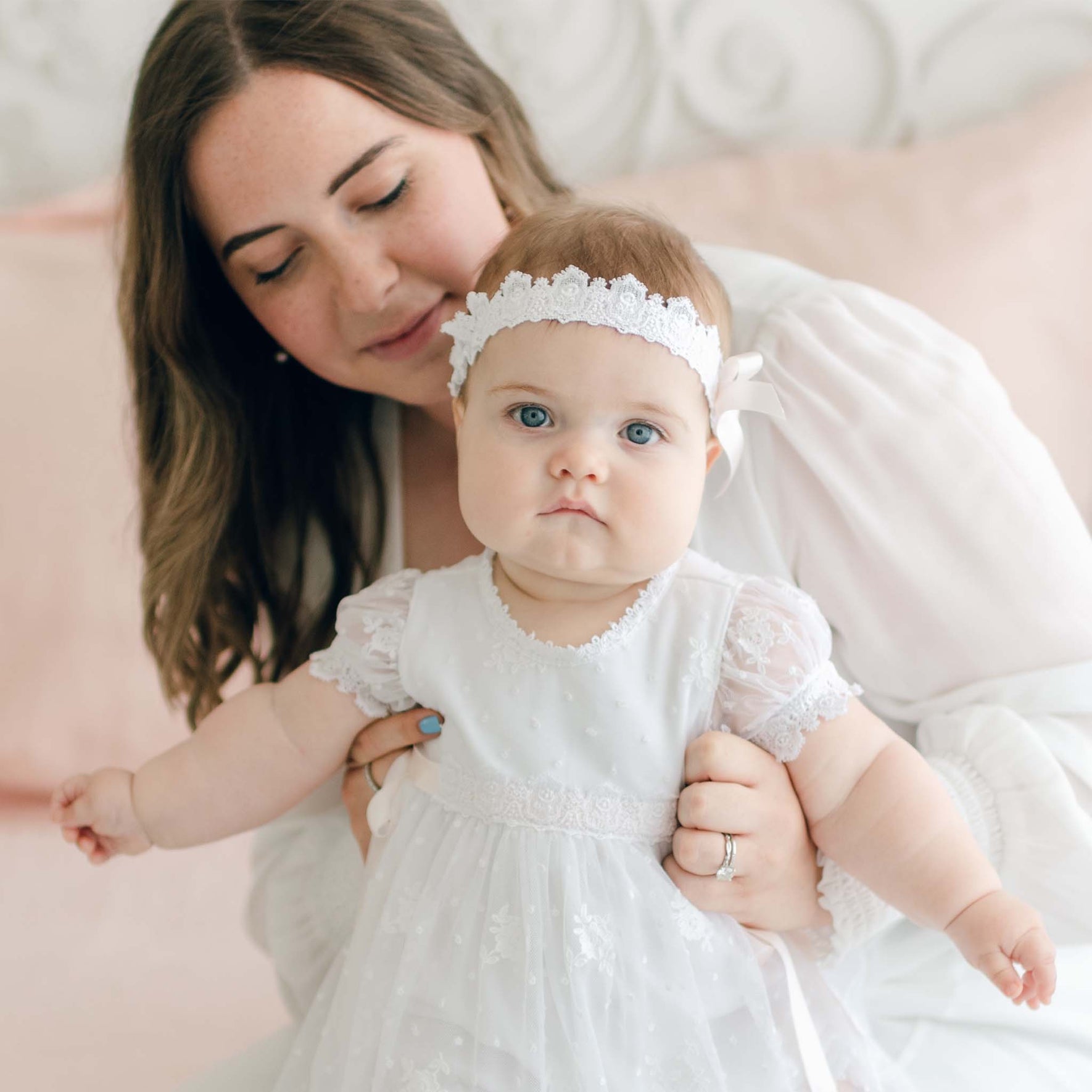 A woman with shoulder-length brown hair holds a baby dressed in a white lace dress adorned with a silk ribbon bow and the Melissa Headband. The baby, with blue eyes and a neutral expression, is being supported by the woman. The background is softly blurred with pink and white tones.