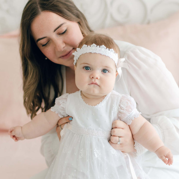 A woman with shoulder-length brown hair holds a baby dressed in a white lace dress adorned with a silk ribbon bow and the Melissa Headband. The baby, with blue eyes and a neutral expression, is being supported by the woman. The background is softly blurred with pink and white tones.