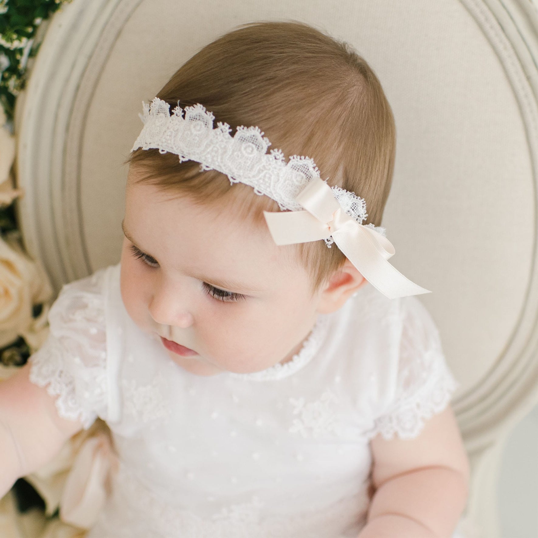 A baby wearing a white Melissa Headband with a pink silk ribbon bow sits on a light-colored chair. The baby is dressed in a white lace dress and gazes off to the side. Roses are partially visible in the background.