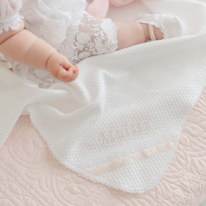 A baby wearing a white lace dress is laying on a quilted textured cotton Melissa Personalized Blanket embroidered with the name "Melissa" in light pink. The baby's left arm and legs are visible. The blanket, handmade in the USA, also has Venice lace trim and a pink ribbon detail.