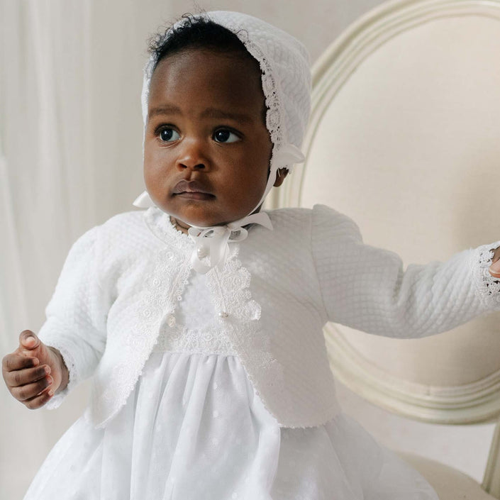 Baby in a white cotton sweater with a bonnet sitting on a chair.