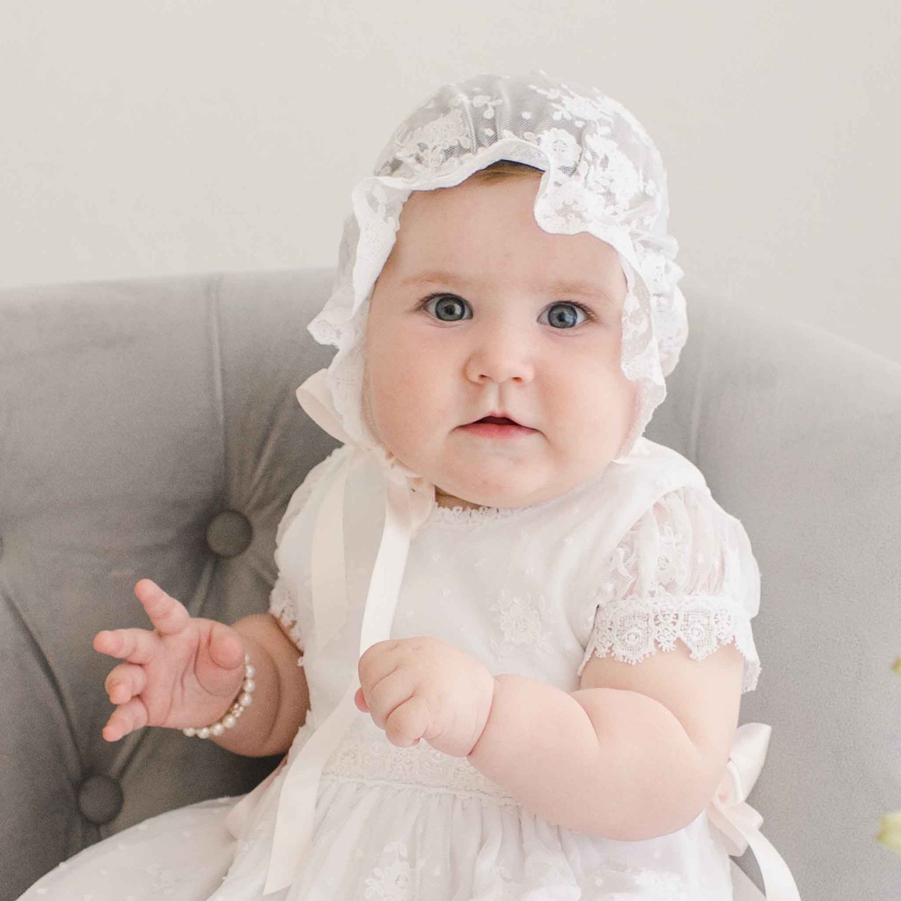 A baby girls sits on a cushioned chair wearing a Melissa Sheer Christening Bonnet and matching baptismal dress. She gazes directly at the camera with a curious expression.