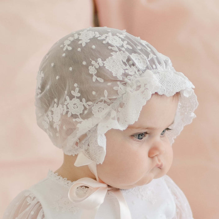 Side profile of a baby girl wearing the Melissa Sheer Christening Bonnet, featuring delicate floral lace and scalloped edges. The bonnet is tied with soft satin ribbons under her chin.