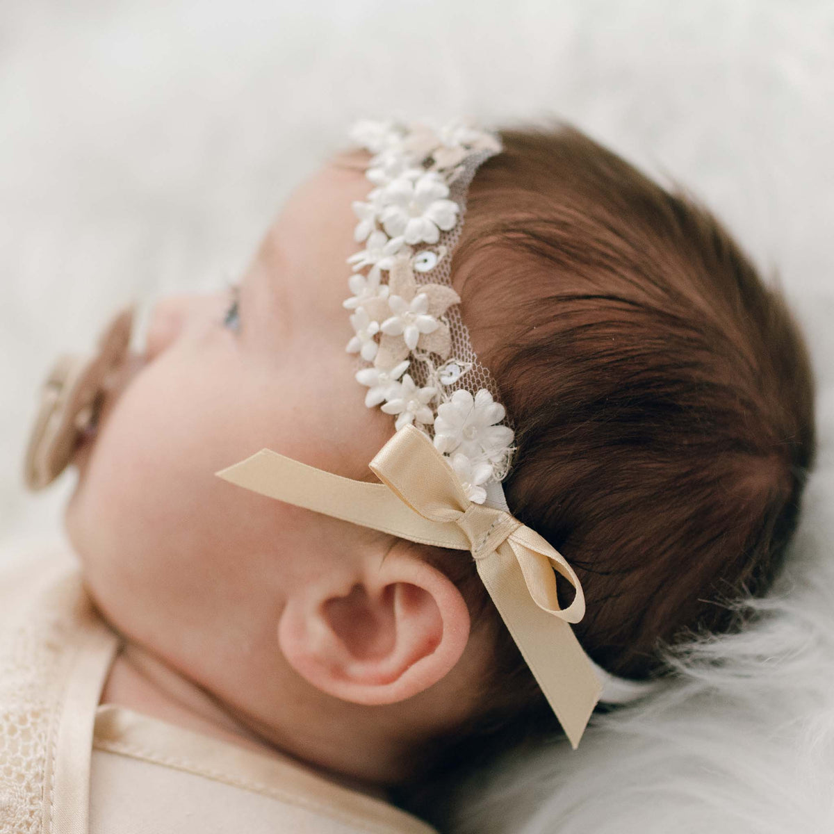 A newborn baby with dark hair wearing the Mia Headband with floral pearlesque beading and a gold silk ribbon bow, lying on a soft white blanket.