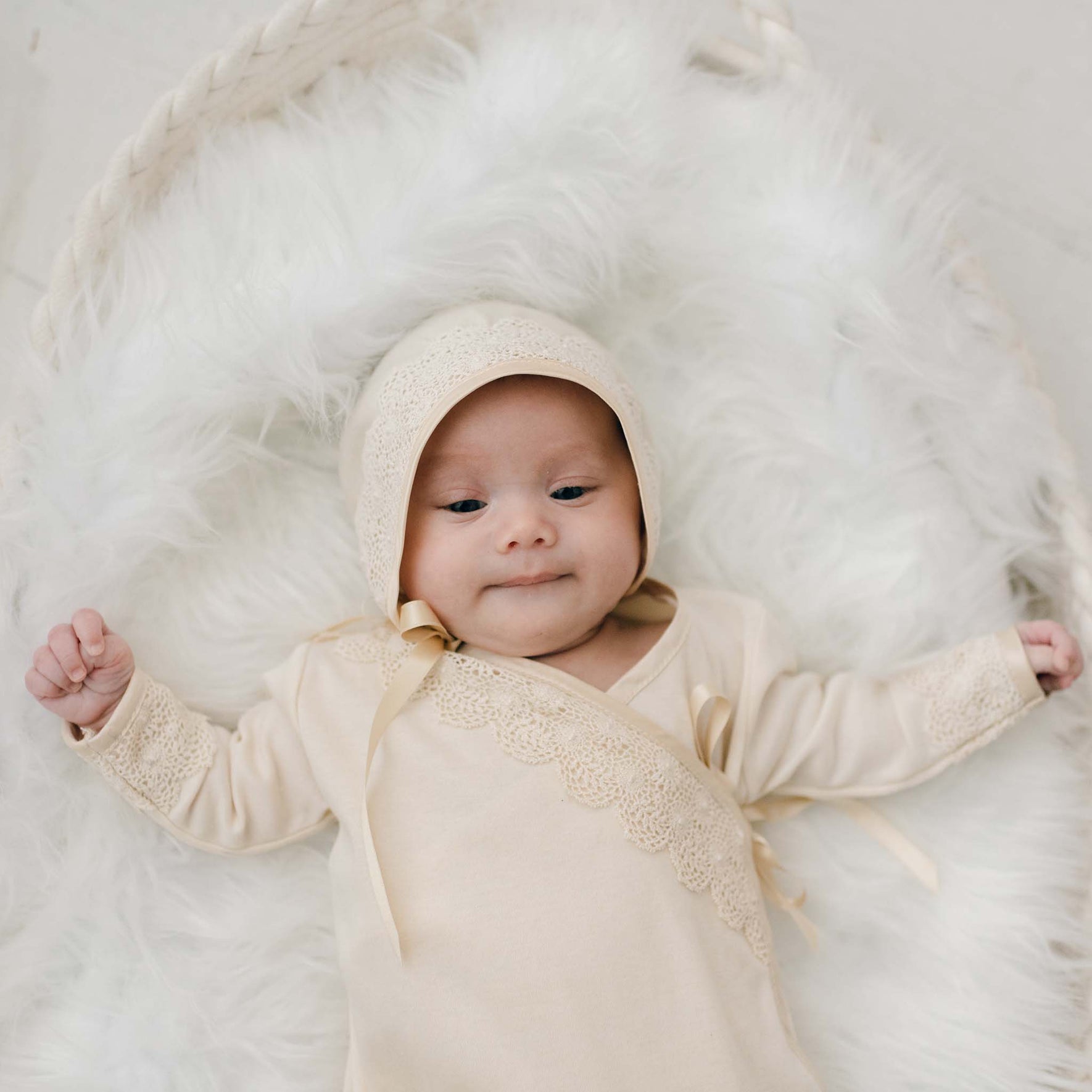 A baby in the Mia Newborn Christening Knot Gown & Bonnet, a cream-colored and vintage-inspired outfit, rests on a fluffy white surface. Made from soft pima cotton, this elegant attire is designed for newborns. The baby gazes up with open eyes and slightly elevated arms in the cozy setting.