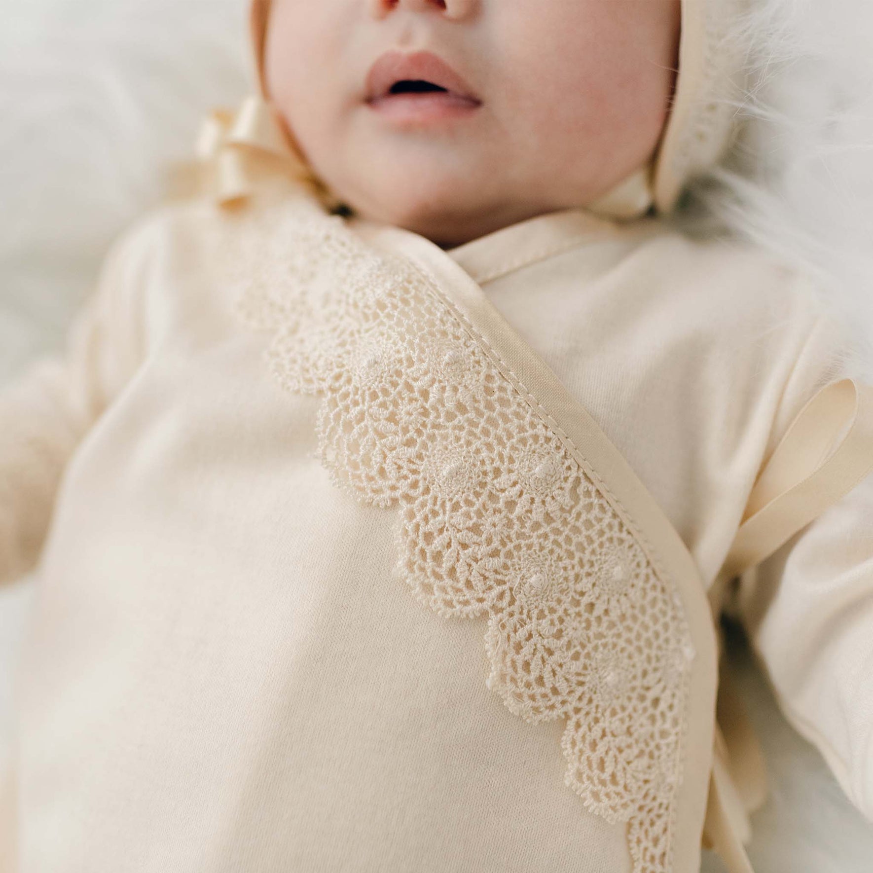 A close-up features a baby in the Mia Newborn Christening Knot Gown & Bonnet, a vintage-inspired beige outfit crafted from soft pima cotton with elegant lace detailing. The look is completed by a matching bonnet. The intricate lace pattern on the front of the gown draws attention against a softly blurred background.