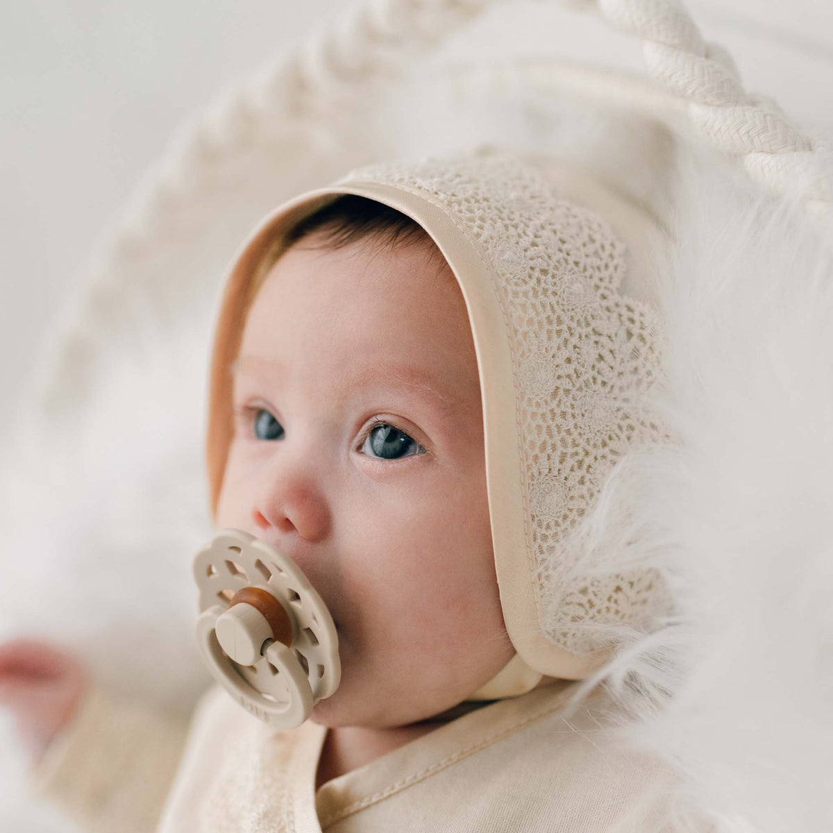 A baby with a pacifier is wearing the Mia Newborn Christening Knot Gown & Bonnet, featuring a vintage-inspired cream-colored lace design. This soft ensemble beautifully highlights the baby's blue eyes as they lie on a plush white surface.