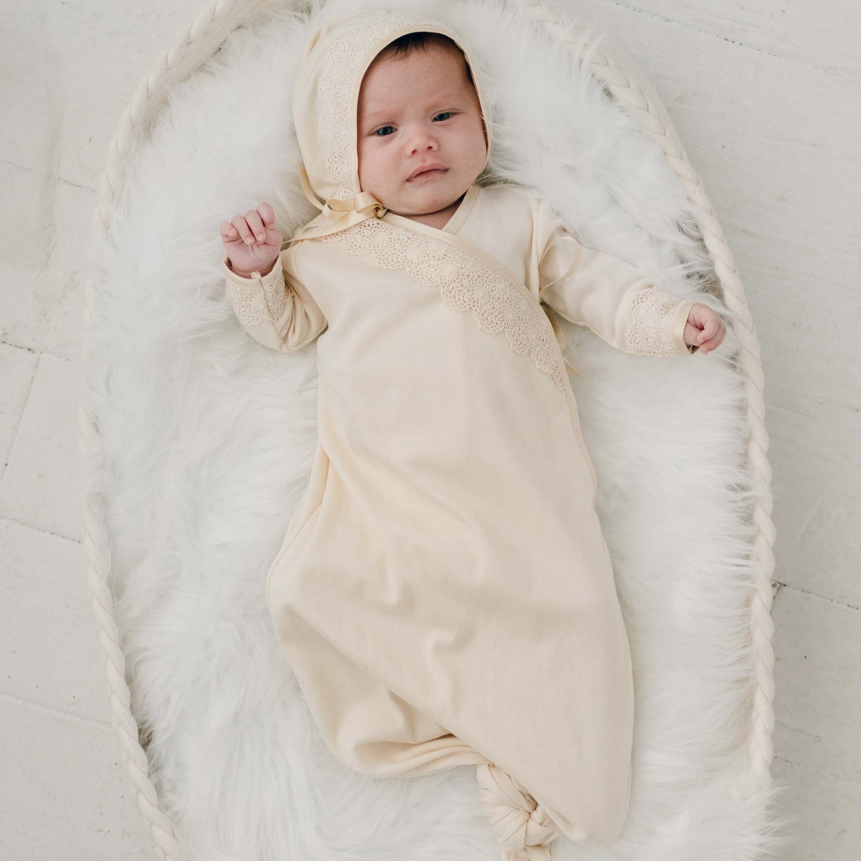 A newborn, dressed in the Mia Newborn Christening Knot Gown & Bonnet, featuring a vintage-inspired cream color and intricate lace details, rests in a cushioned basket lined with white fur. The scene is set on a light surface, with the baby gazing upwards serenely.