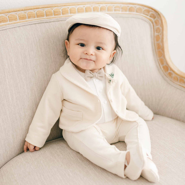 A baby in the Milo Tan 3-Piece Suit sits on an elegant couch, wearing a tan jacket, bow tie, and pants with matching cap and booties.