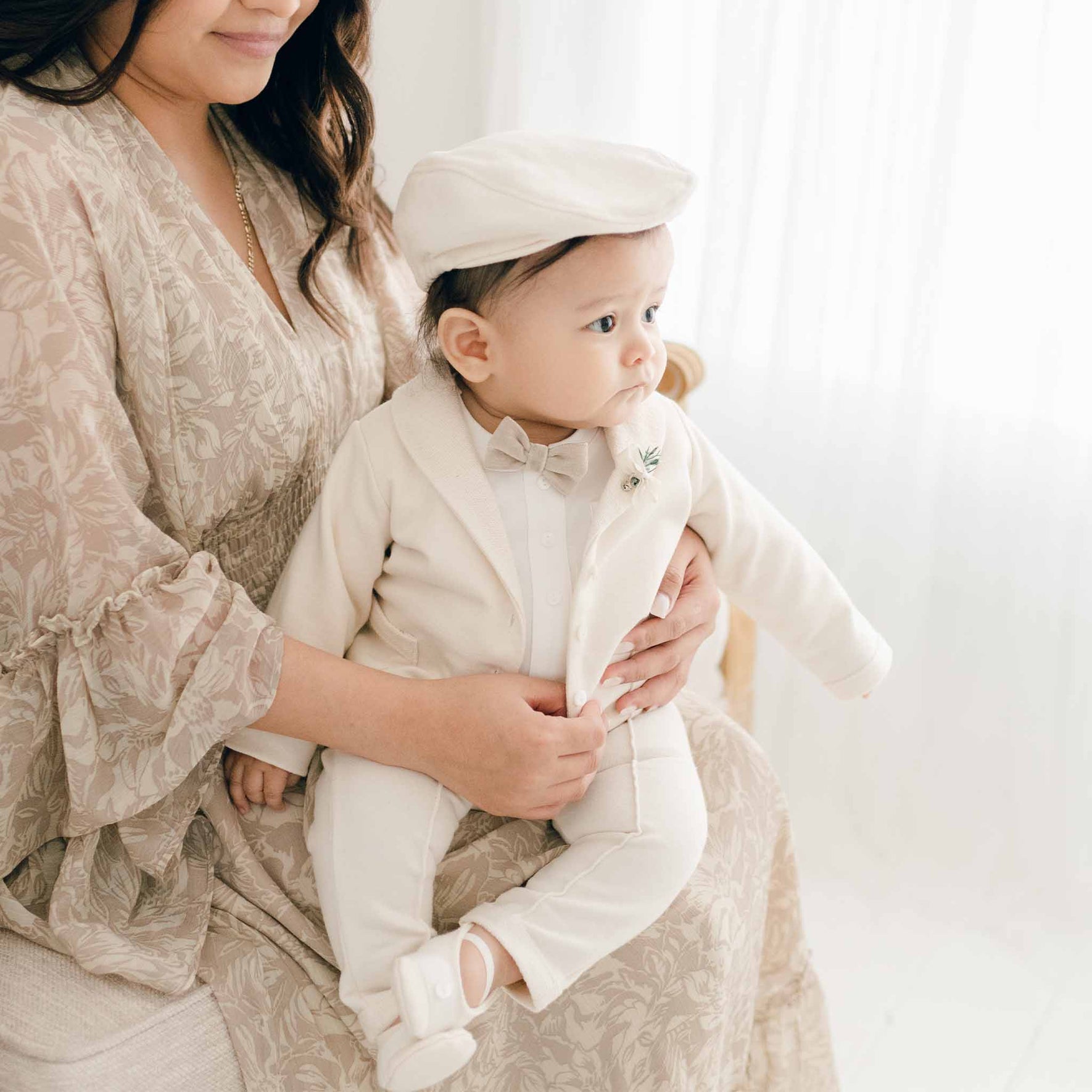 A baby in the Milo Tan 3-Piece Suit sits on a woman’s lap, facing sideways, dressed in a jacket, bow tie, and cap.
