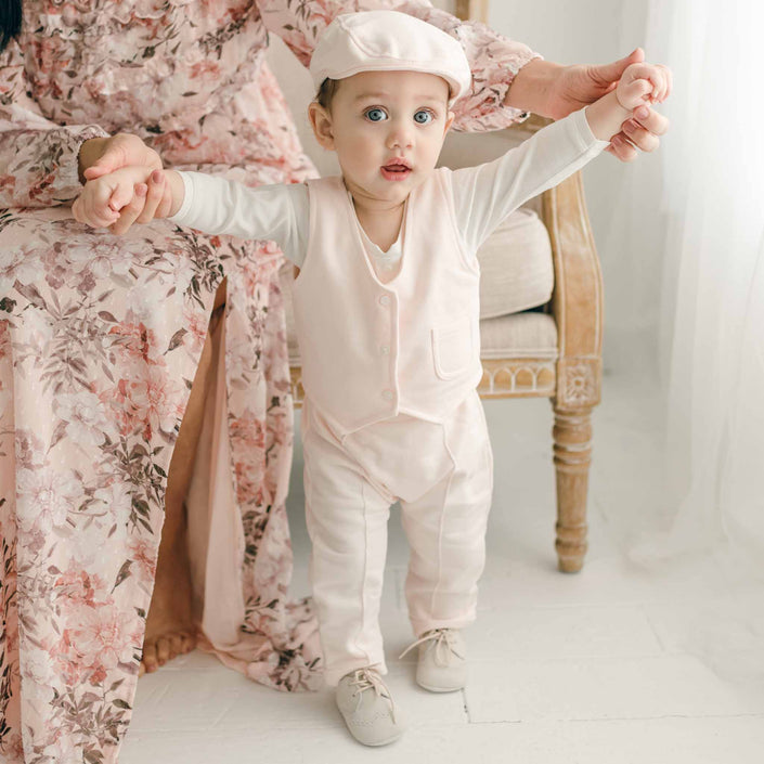 A baby in the Milo Blush Vest, standing with assistance, wearing a coordinating cap and pants.