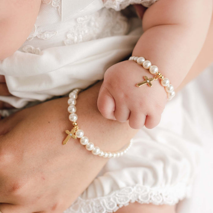 A close-up lifestyle shot of a baby's wrist and ankle adorned with matching pearl bead bracelets and anklets, each featuring a small gold cross charm, worn with a white lace-trimmed outfit.
