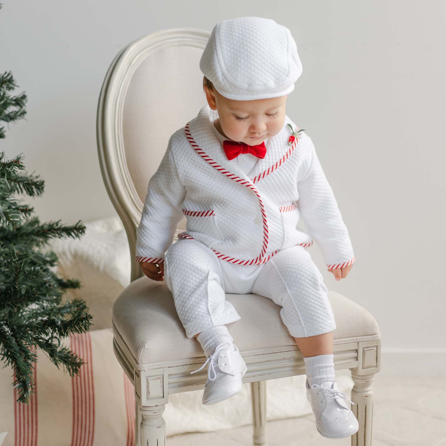 Baby in a white outfit with red trim sitting on a chair next to a Christmas tree.