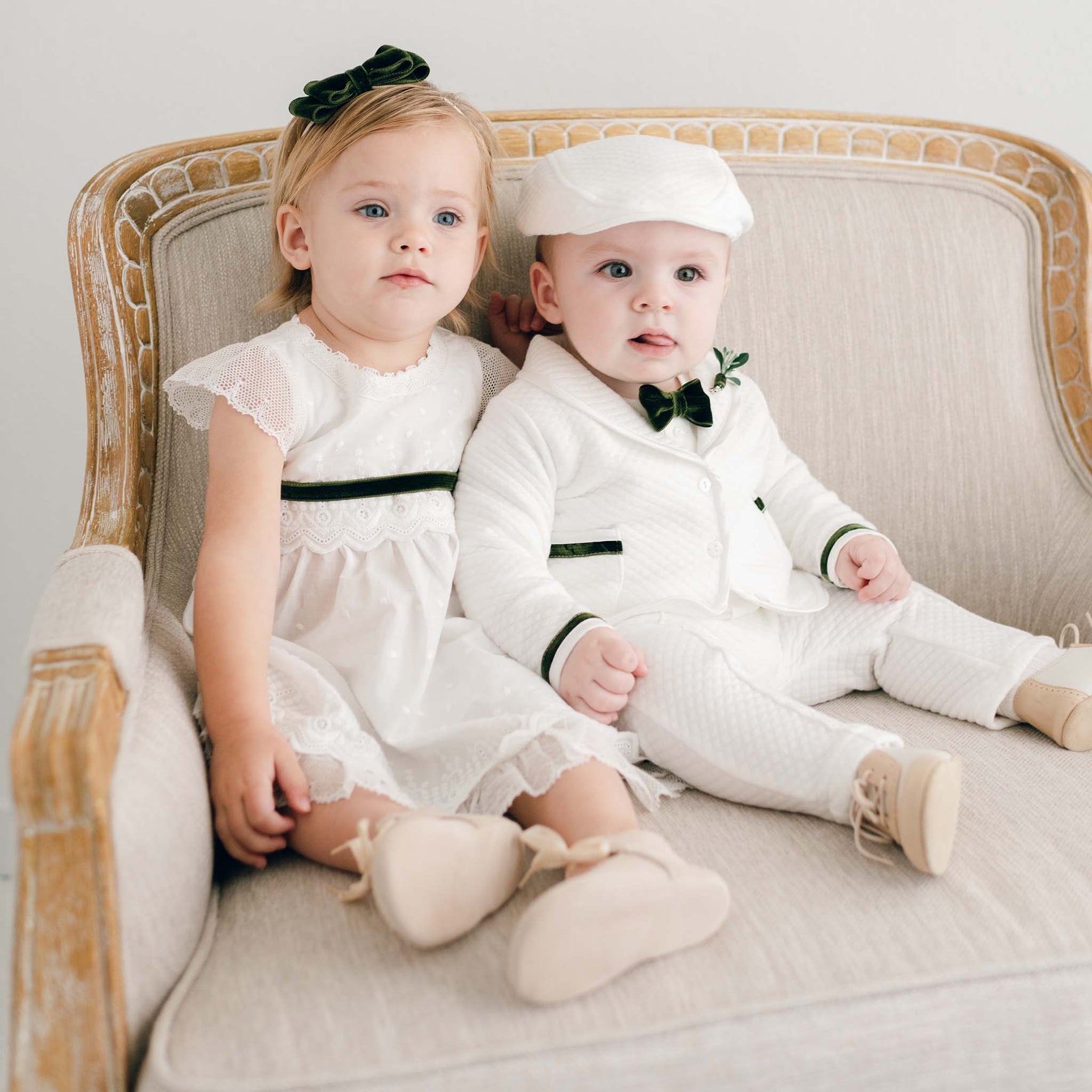 Two toddlers are seated on a beige upholstered armchair. The girl is dressed in a white dress with lace sleeves and a green headband, while the boy is sporting an adorable Christmas outfit—a Noah 3-Piece Suit with green trim, complete with a matching cap and bow tie. Both are looking ahead.