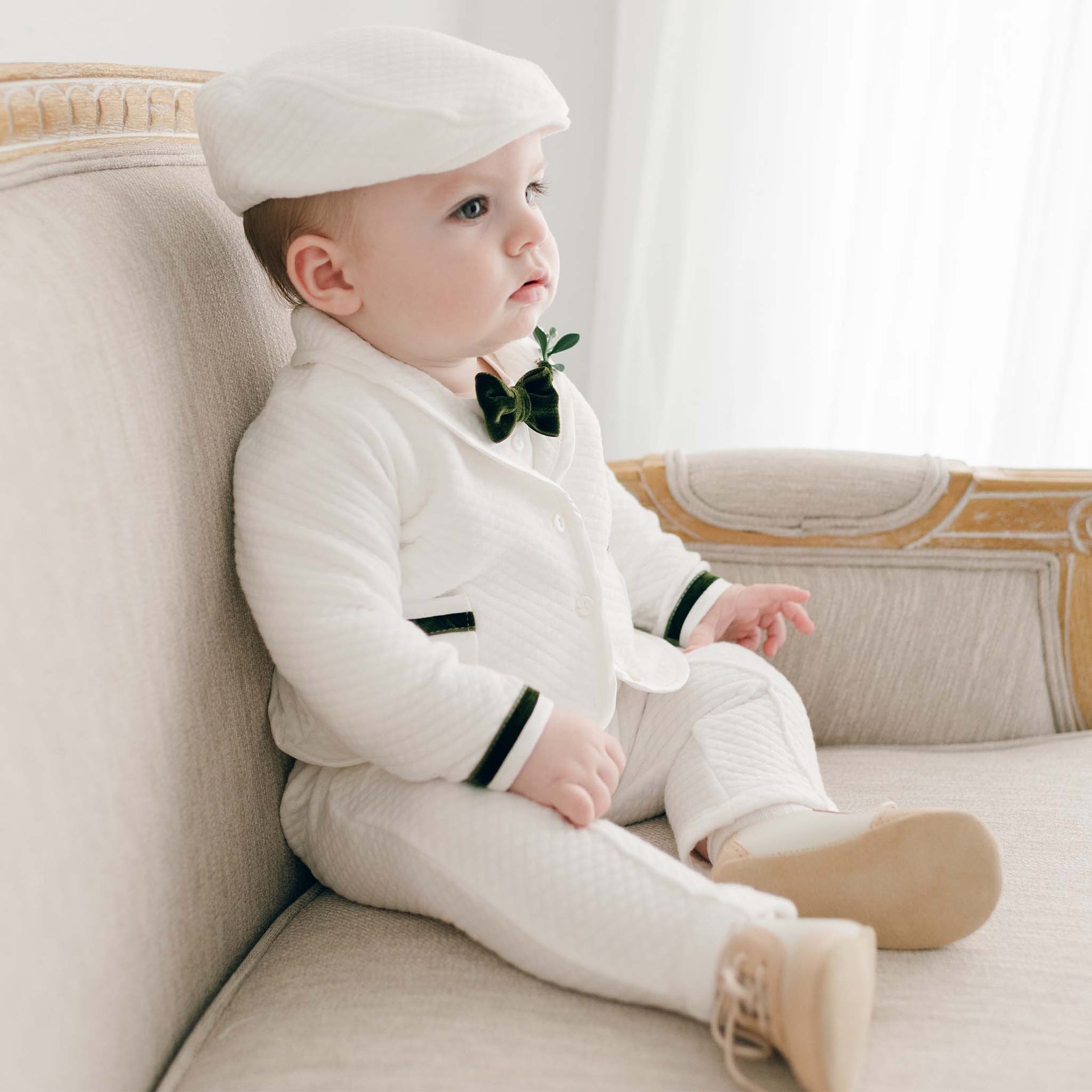A baby is sitting on a beige couch, looking dapper in the Noah 3-Piece Suit with green trim, a matching white hat, and a black bow tie. The calm expression on his face complements the softly lit background, making it perfect for a Christmas outfit.