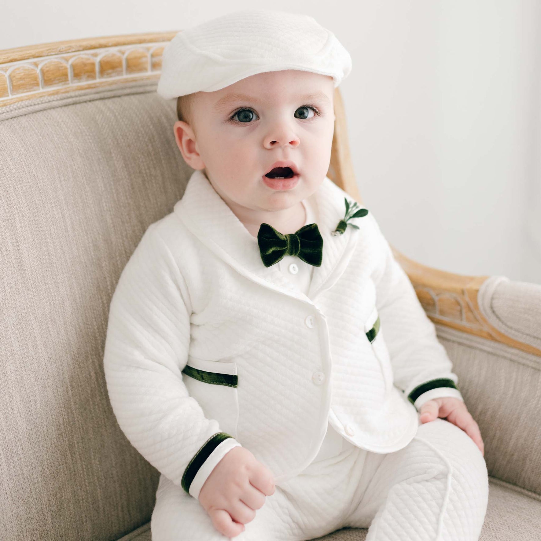 A baby, dressed in the Noah 3-Piece Suit with green trim, sits on a beige armchair. The quilted cotton outfit is complete with a bow tie and matching cap, giving the little one a charming appearance. With a surprised expression, the baby gazes forward.