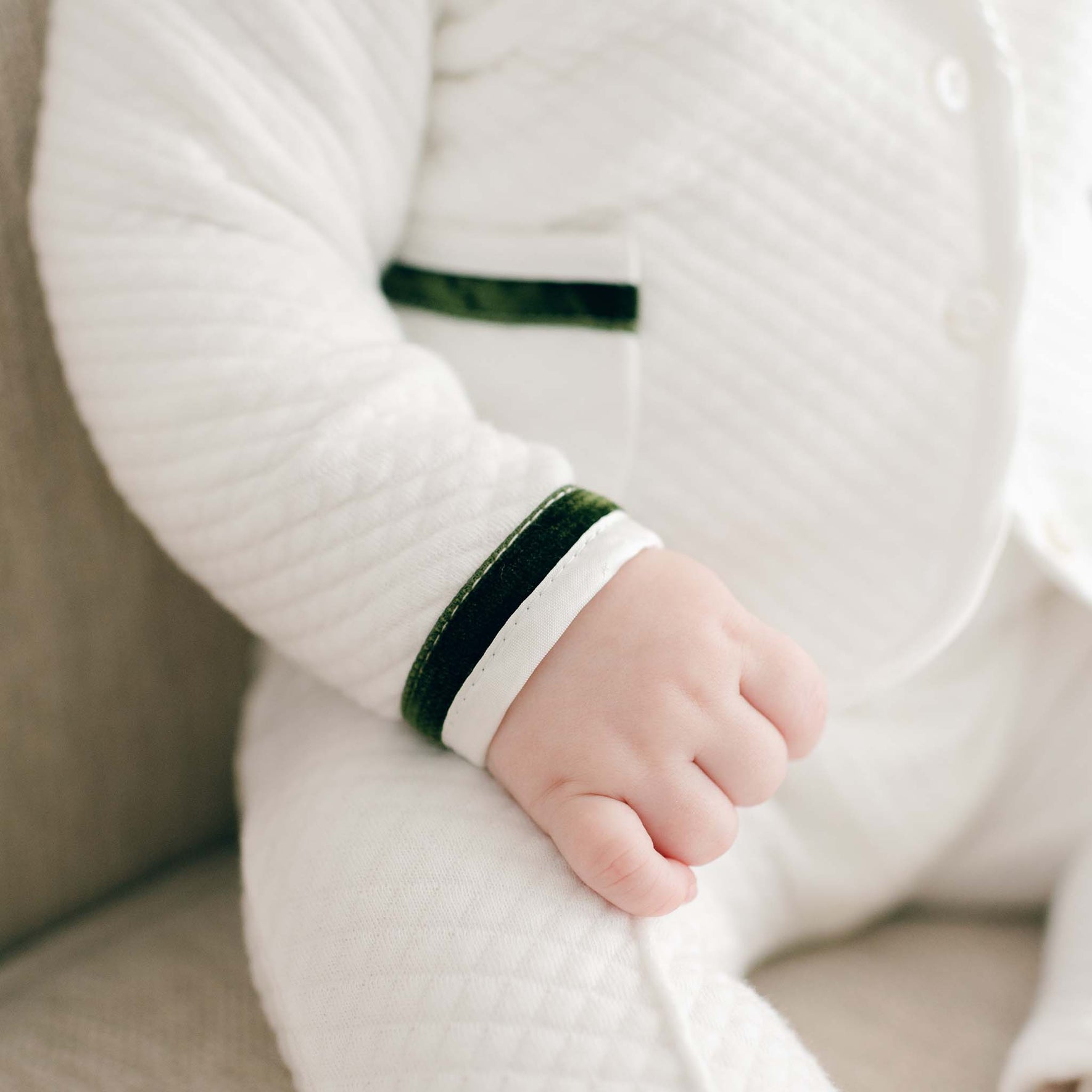 A baby wearing the Noah 3-Piece Suit with textured white quilted cotton and green trim is sitting. The Christmas outfit is adorned with button details and a visible pocket. The baby's hand rests relaxed on their lap while seated on a light-colored surface.