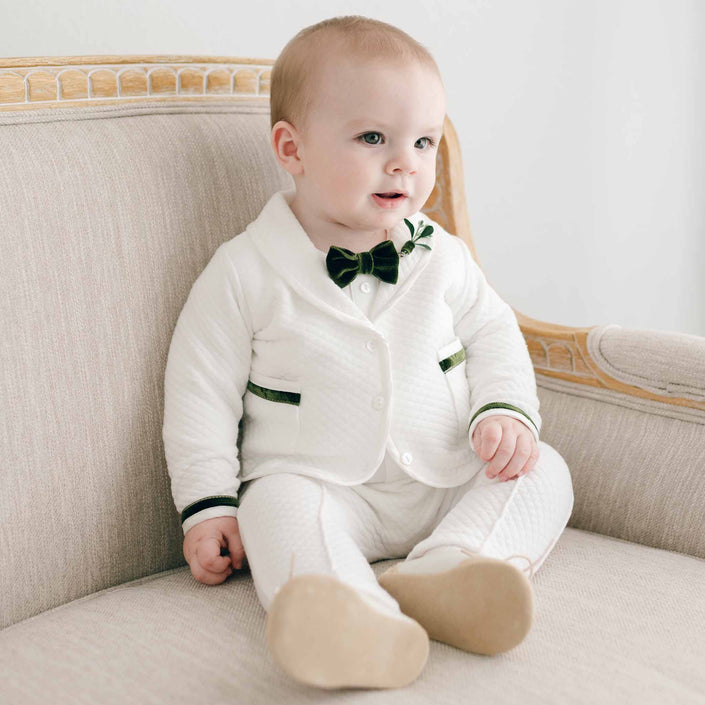 A baby boy in the "Noah 3-Piece Suit | Green Trim," featuring a formal white suit with green accents and a bow tie, sits on a beige sofa with wooden edges and a cushioned back. Wearing his Christmas outfit, he looks to the side with a neutral expression, perfectly complementing the cozy setting.