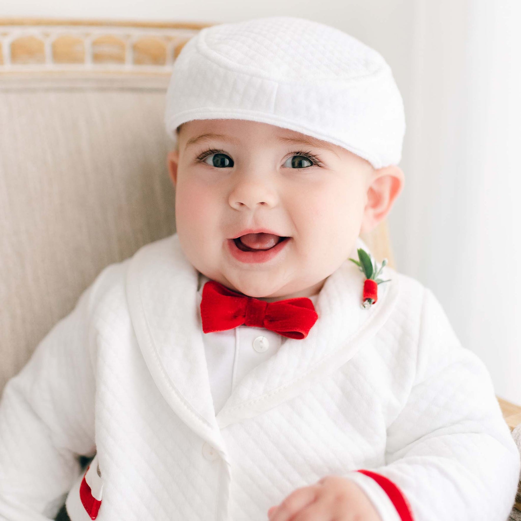 A baby boy is sitting on a chair, dressed in the Noah 3-Piece Suit with red trim, which includes a charming white outfit and a matching cap. He sports a red velvet ribbon bow tie and has a small flower pinned to the lapel, beaming with an open-mouthed smile, ready for Christmas parties.