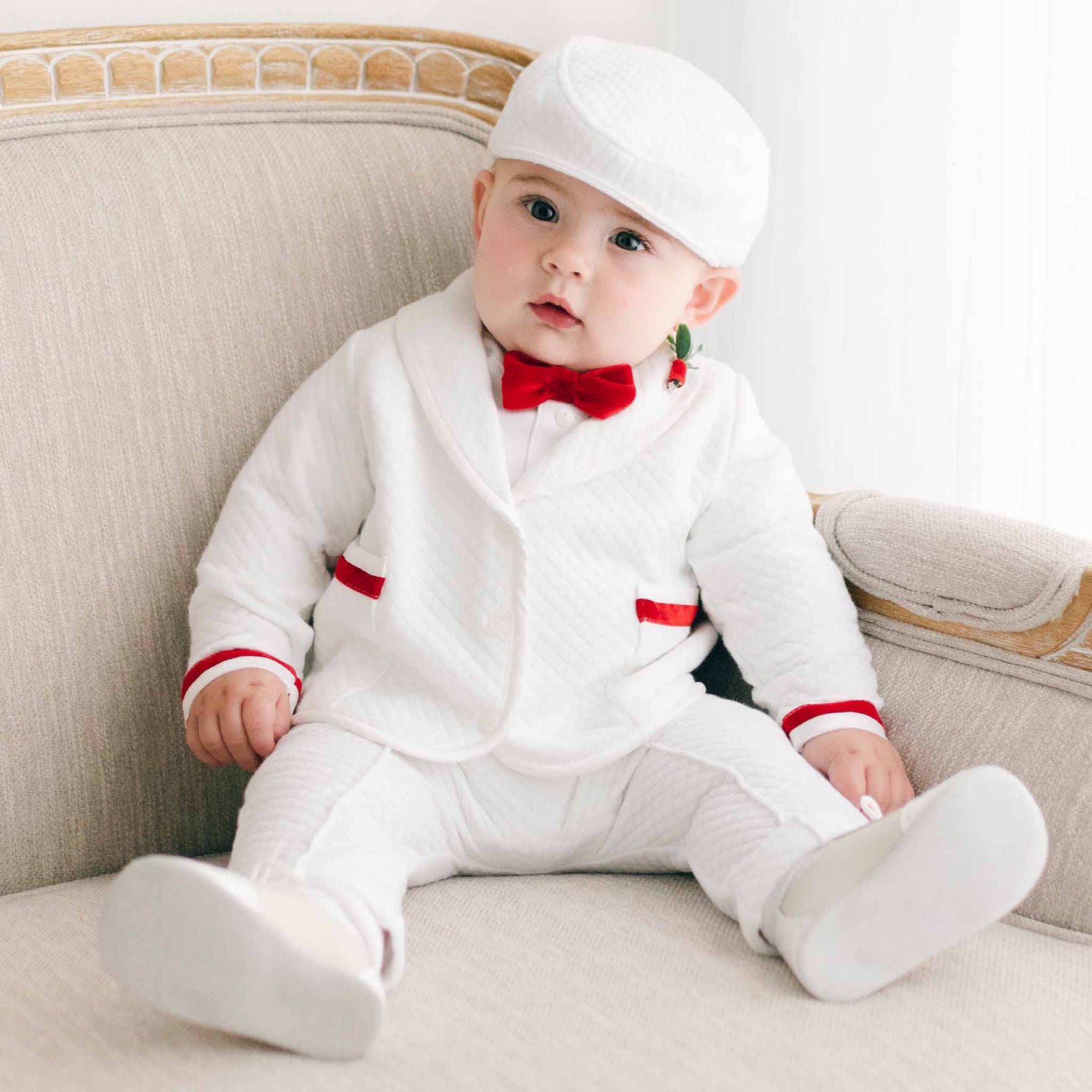 A baby boy dressed in the Noah 3-Piece Suit with red trim sits on a beige couch, making it an ideal choice for Christmas parties. The outfit features a crisp jacket with red-trimmed pockets, a red velvet ribbon bow tie, and is completed with a matching white hat. A small decorative toy hangs from one ear for an added festive touch.