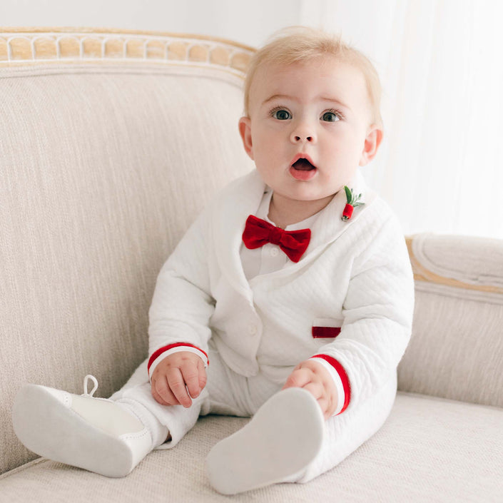 Dressed in the Noah 3-Piece Suit with red trim, a baby boy sits on a beige sofa. The suit features charming white fabric accented by a red bow tie and velvet ribbon. With wide eyes and a slightly open mouth, he's all set for Christmas parties. His festive look is completed with white shoes.