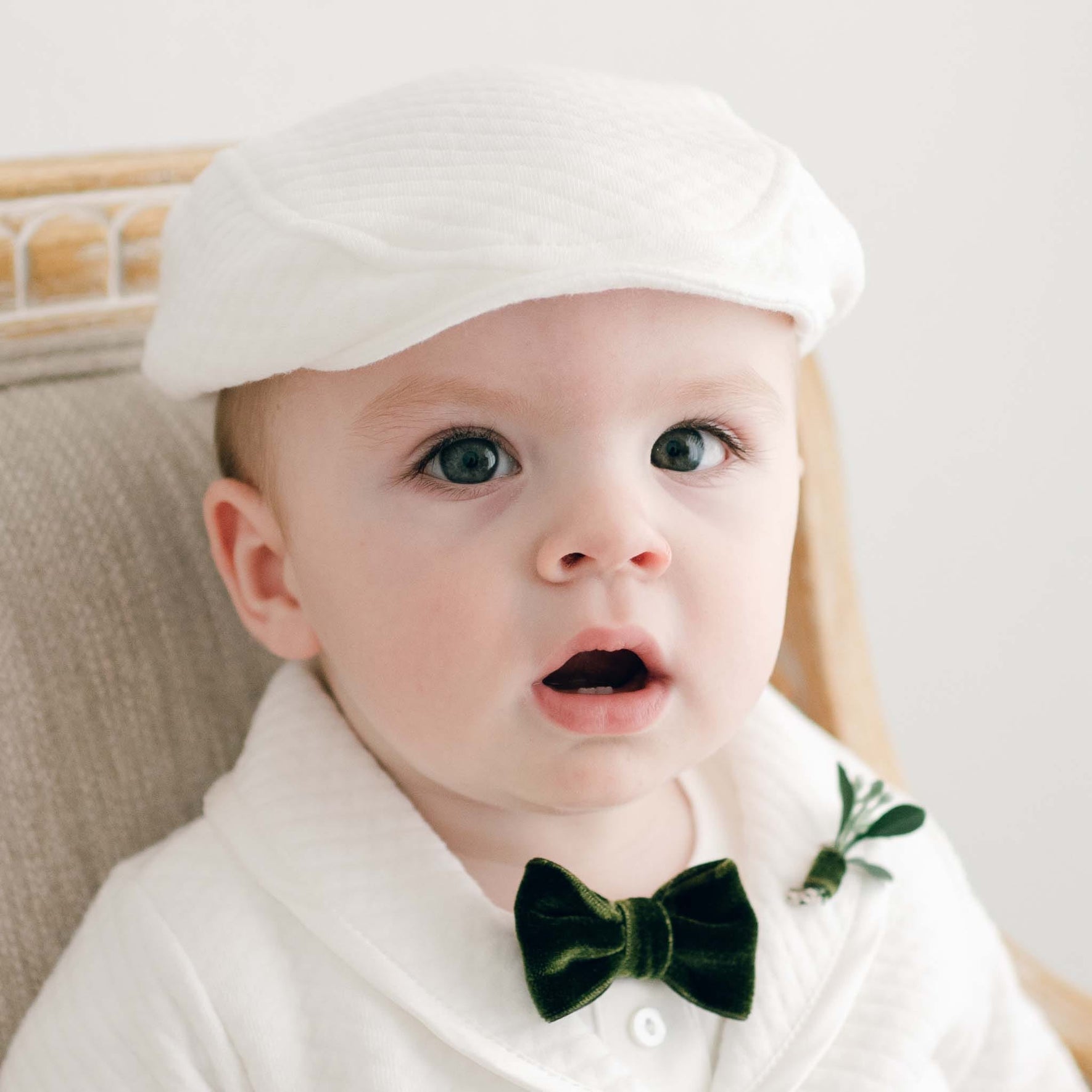 A baby wearing a white cap, a white jacket, and the Noah Green Bow Tie & Boutonniere sits on a chair. The holiday accessory adds charm as the baby gazes upward with an open mouth. The soft, neutral background highlights the child’s attire, handmade in the USA.