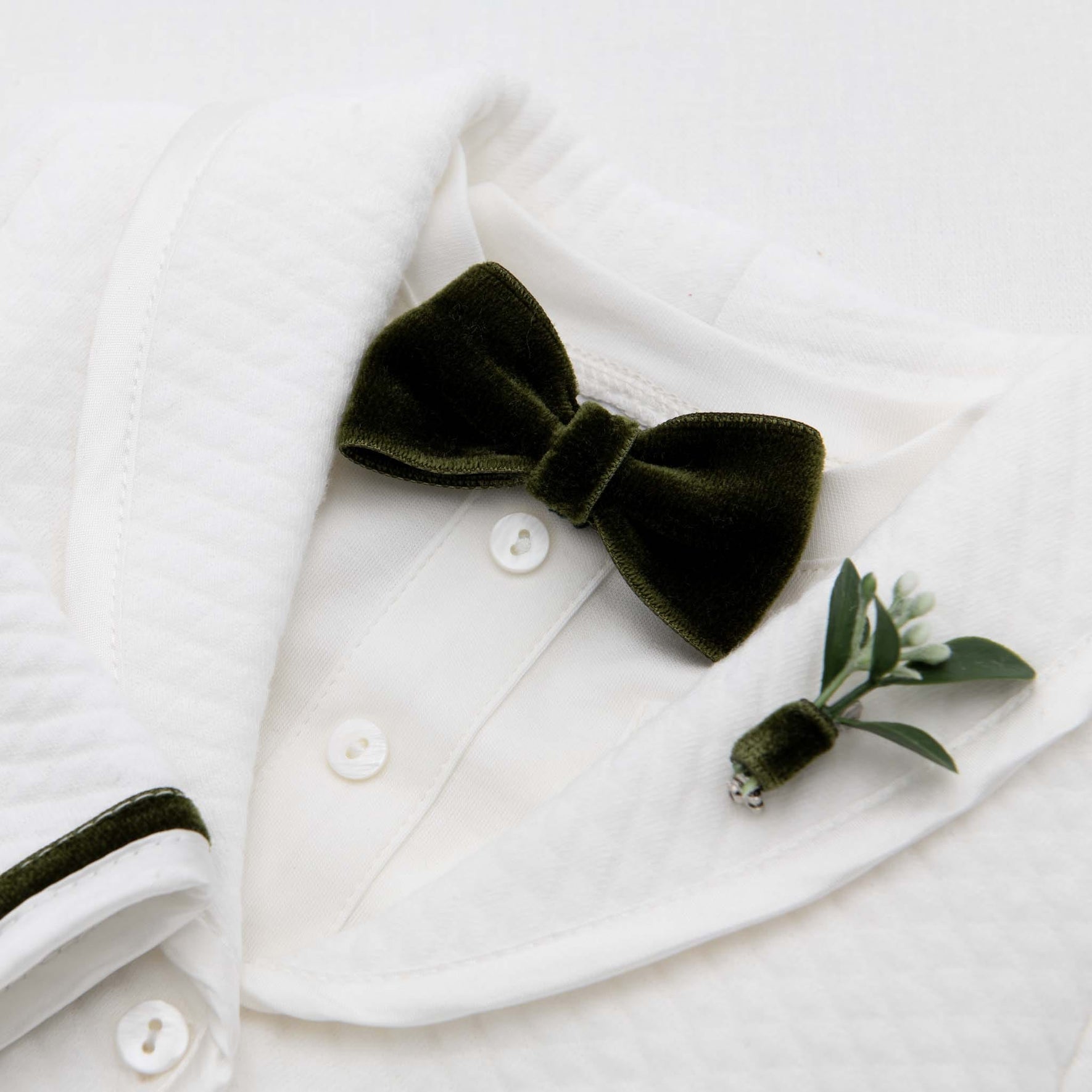 The Noah Green Bow Tie & Boutonniere set showcases a luxurious dark green velvet bow tie, handmade in the USA, elegantly paired with a small boutonniere of greenery and a white flower bud, pinned on the left lapel of a textured white dress shirt. It's the perfect holiday accessory for any elegant occasion.