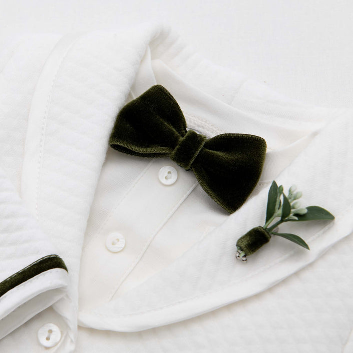 The Noah Green Bow Tie & Boutonniere set showcases a luxurious dark green velvet bow tie, handmade in the USA, elegantly paired with a small boutonniere of greenery and a white flower bud, pinned on the left lapel of a textured white dress shirt. It's the perfect holiday accessory for any elegant occasion.