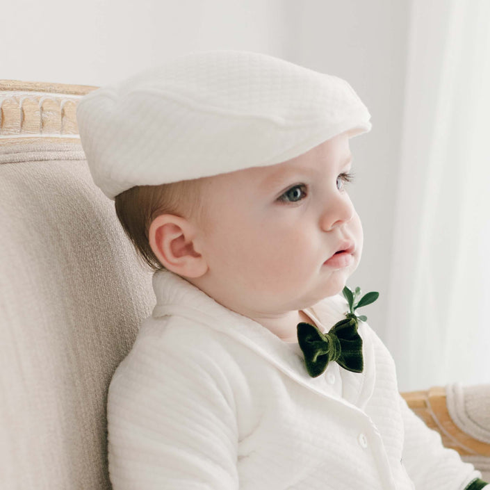 A baby is sitting on a plush chair, dressed in the Noah Ivory Newsboy Cap along with a coordinating white outfit. This handmade ensemble from the USA includes a small dark green bow tie accented with a sprig of greenery. The baby gazes to the side in a brightly lit setting.