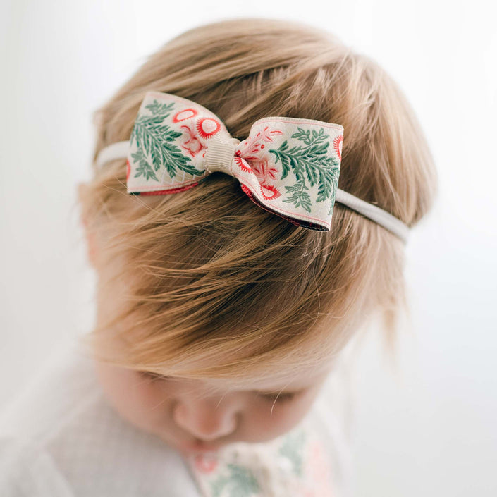 A young child with light brown hair is wearing a Noelle Headband, which includes a decorative bow crafted from holiday floral ribbon in green and pink. The bow is handmade in the USA. The child is gazing downward with a softly blurred background behind them.