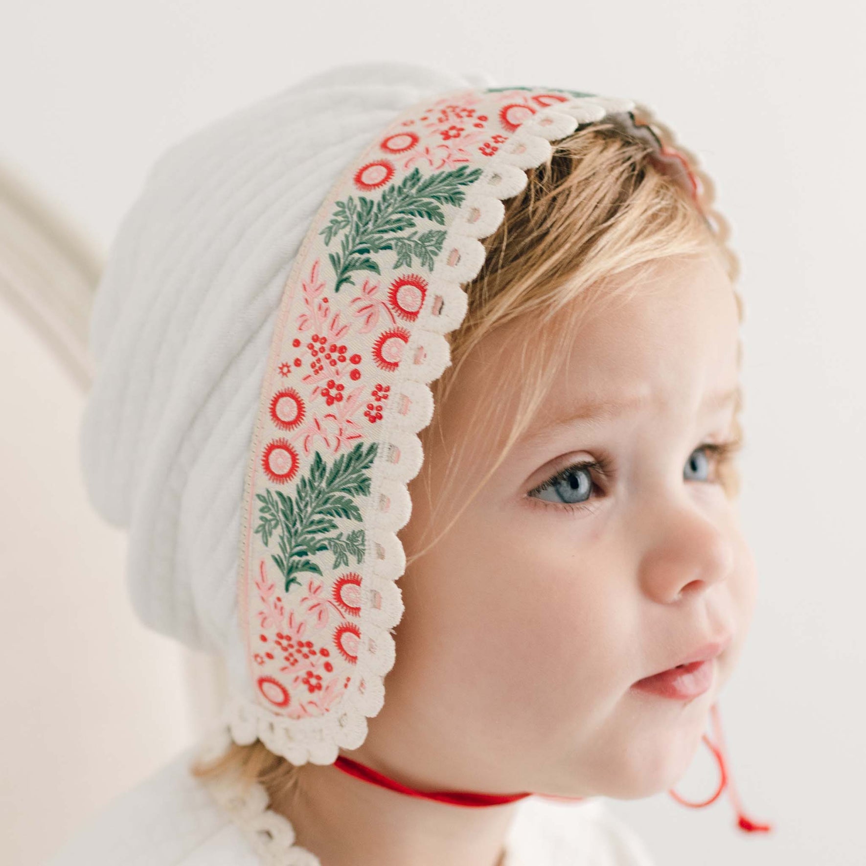 A young child wearing the Noelle Quilt Bonnet, featuring red and green floral patterns with lace trim, beautifully frames their blonde hair and blue eyes as they gaze slightly to the right. The handmade in the USA bonnet stands out against a soft, neutral backdrop, ideal for the holiday season.