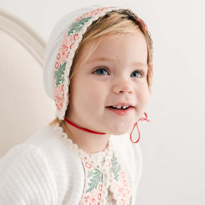 A young child with blue eyes smiles while wearing the Noelle Quilt Bonnet, a holiday season bonnet in white adorned with red and green floral patterns and a red string. The child is dressed in a matching outfit featuring floral embroidery, creating a charming look. This handmade bonnet from the USA beautifully stands out against the neutral light background.