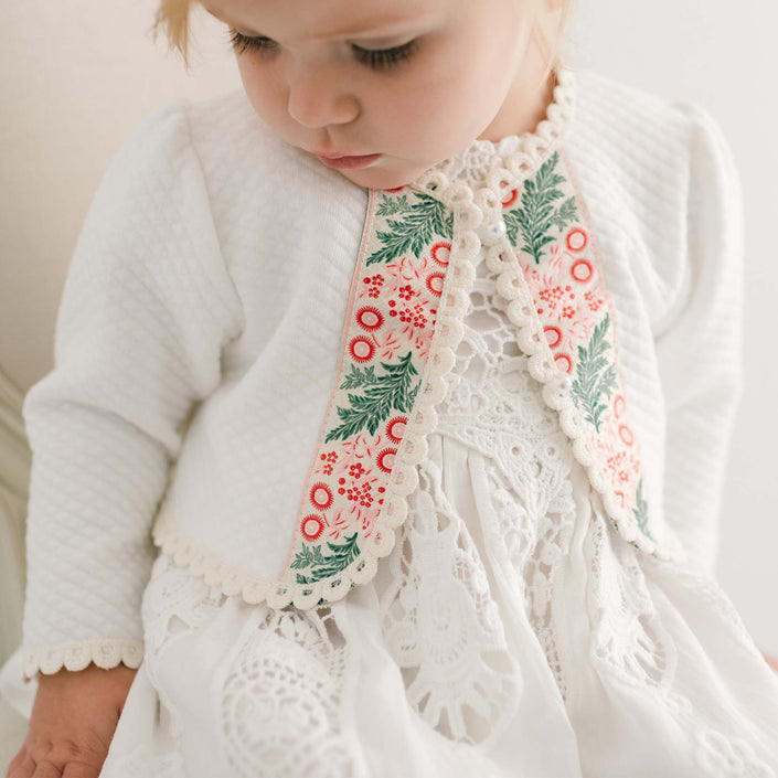 A toddler wearing the Noelle Quilted Cotton Sweater, featuring an ivory quilted cotton dress with ecru edge lace and a matching jacket, ideal for a holiday outfit. The jacket is adorned with a floral pattern in green and red on the trim. The child looks down, set against a softly lit neutral background.