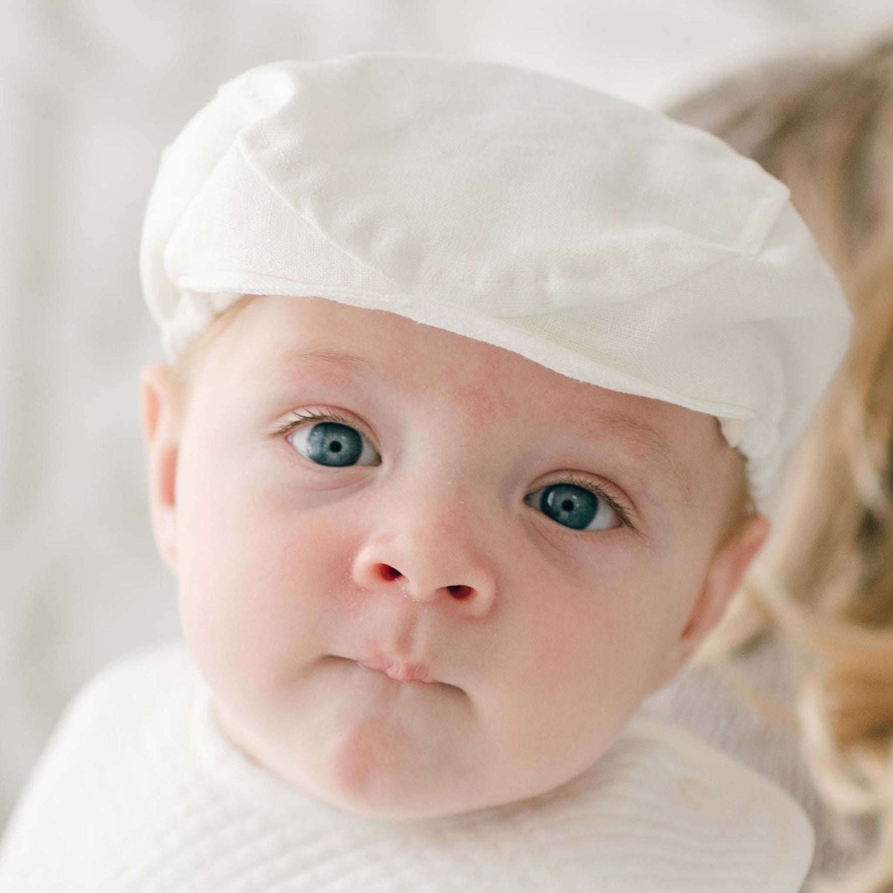 A baby wearing a white cap and a white shirt stares directly into the camera. The baby has blue eyes and slightly pursed lips. The background is softly blurred, highlighting the baby's face in an adorable Oliver Linen Newsboy Cap, handmade in the USA.