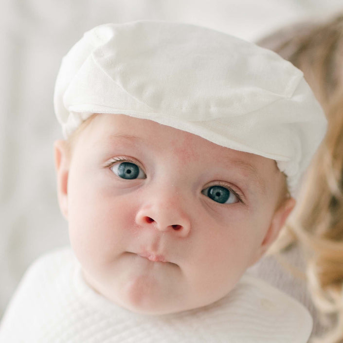 A baby wearing a white cap and a white shirt stares directly into the camera. The baby has blue eyes and slightly pursed lips. The background is softly blurred, highlighting the baby's face in an adorable Oliver Linen Newsboy Cap, handmade in the USA.