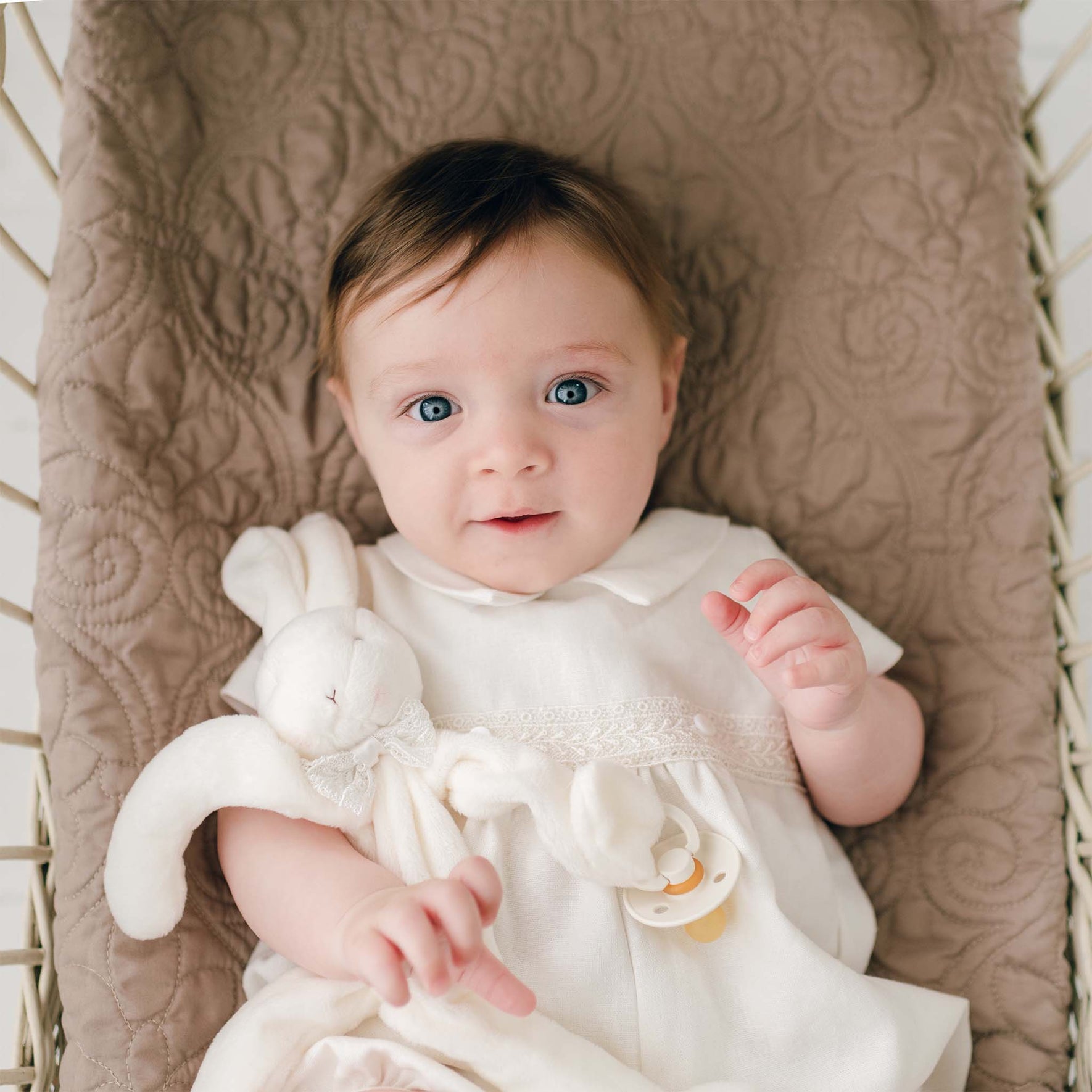 A baby with light brown hair and blue eyes lies on a quilted beige blanket, wearing the Oliver Romper in white. The baby holds an Oliver Silly Bunny Buddy | Pacifier Holder made of soft white velour and has a baby-safe pacifier clip attached to its hand. The baby has a calm, curious expression.