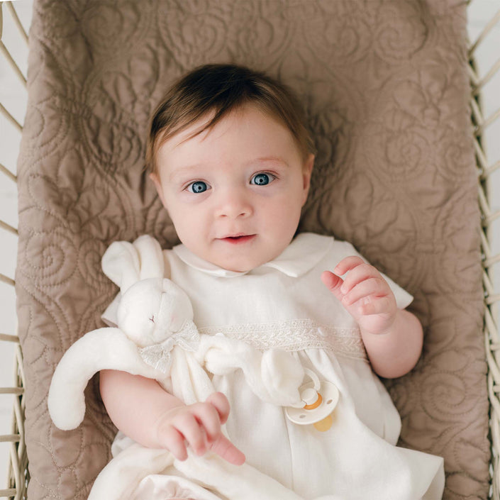 A baby with light brown hair and blue eyes lies on a quilted beige blanket, wearing the Oliver Romper in white. The baby holds an Oliver Silly Bunny Buddy | Pacifier Holder made of soft white velour and has a baby-safe pacifier clip attached to its hand. The baby has a calm, curious expression.