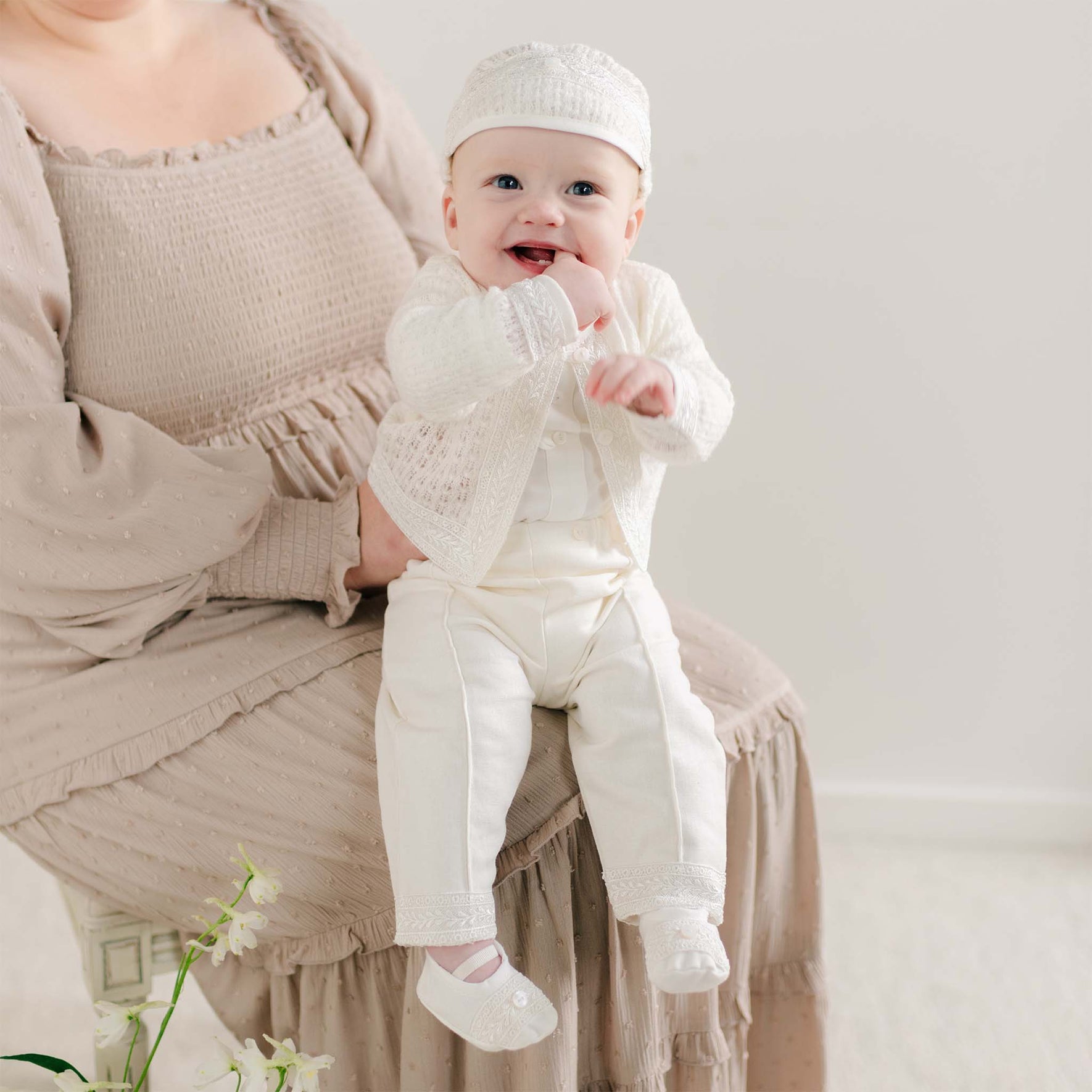 A baby wearing the Oliver Sweater Pants Suit in ivory, sitting on a parent’s lap, with fine knit fabric and lace details on the sweater and pants.