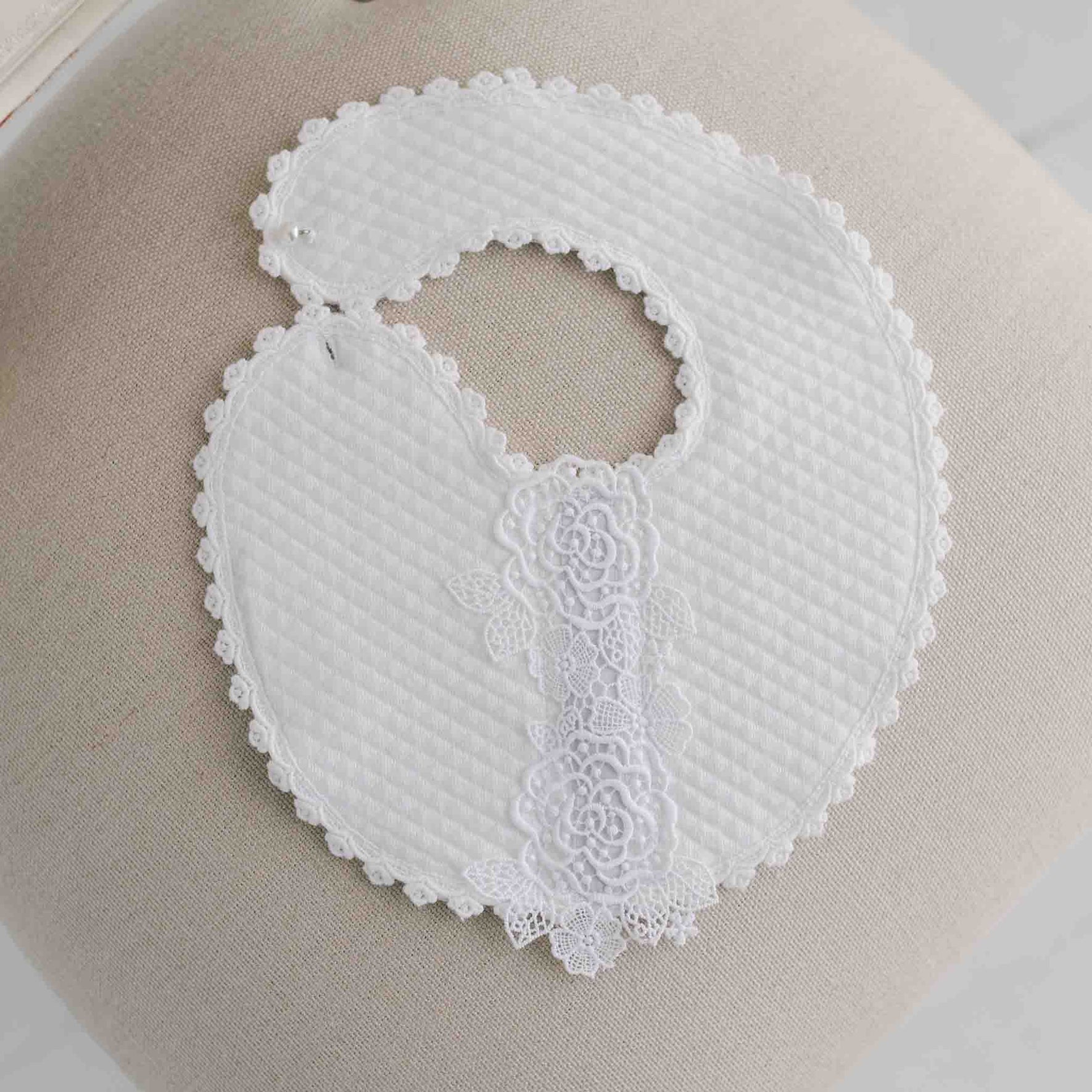 The Olivia Bib, featuring a white, round design ideal for a baptism outfit, is displayed on a beige surface. It boasts scalloped edges and textured fabric with an elegant floral lace design running down the center.